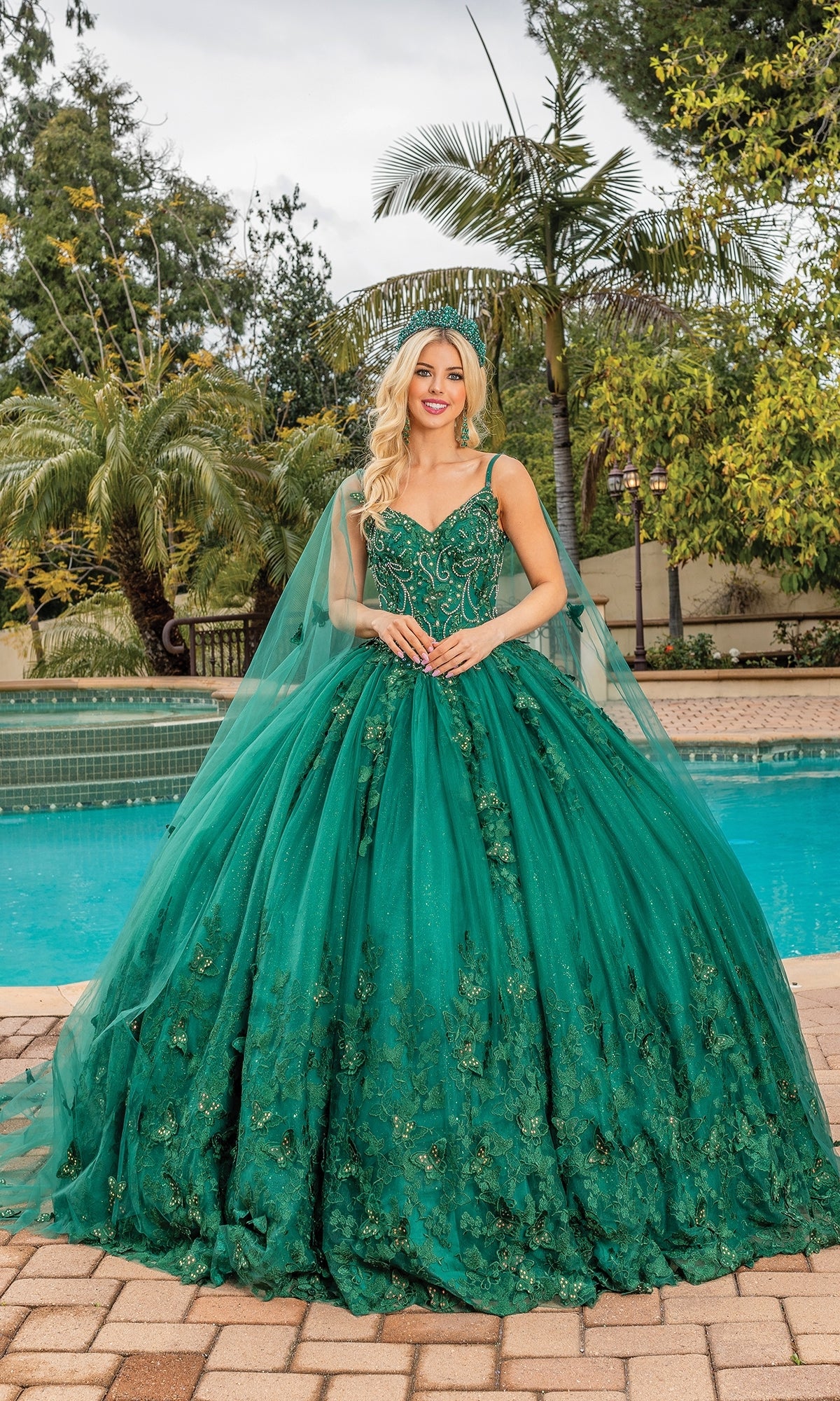 A woman stands by a pool in Quinceanera Dress 1746 By Dancing Queen, featuring a sweetheart neckline, intricate embroidery, floral details, and a long tulle train. Palm trees and lush greenery provide a stunning backdrop.